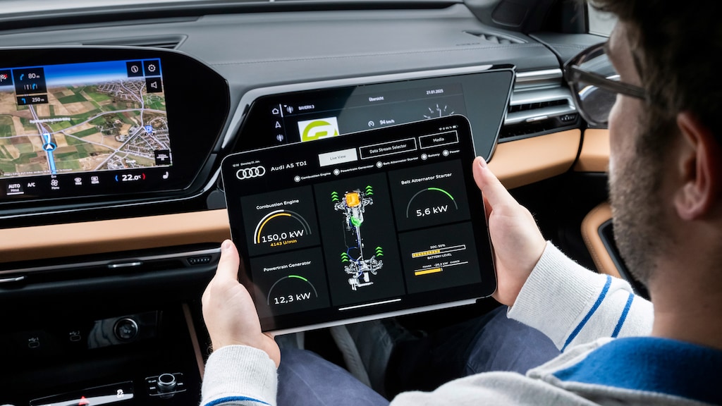 View of a dashboard with monitors. One person is sitting in the passenger seat and holding a tablet on which various graphics can be seen.