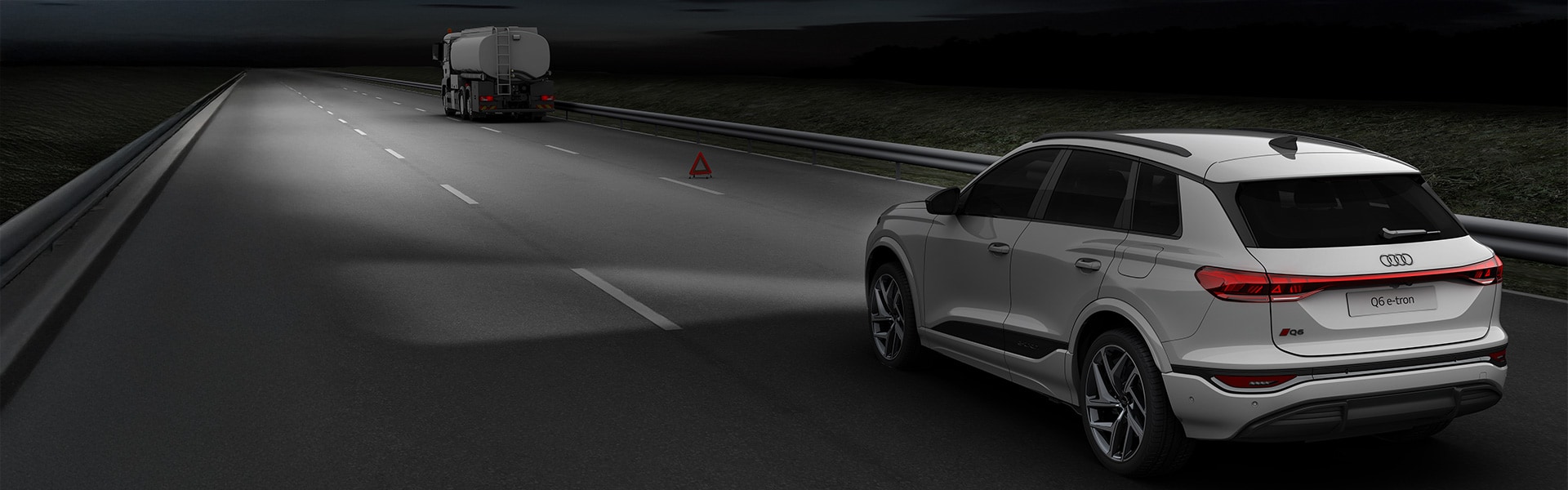 Illustration of a hazardous situation. An Audi model drives towards a lorry in the dark, which is standing on the hard shoulder with a warning triangle.