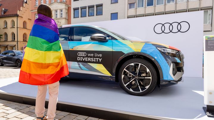 Eine Person steht mit einer umgelegten Pride Flagge vor dem Diversity Audi
